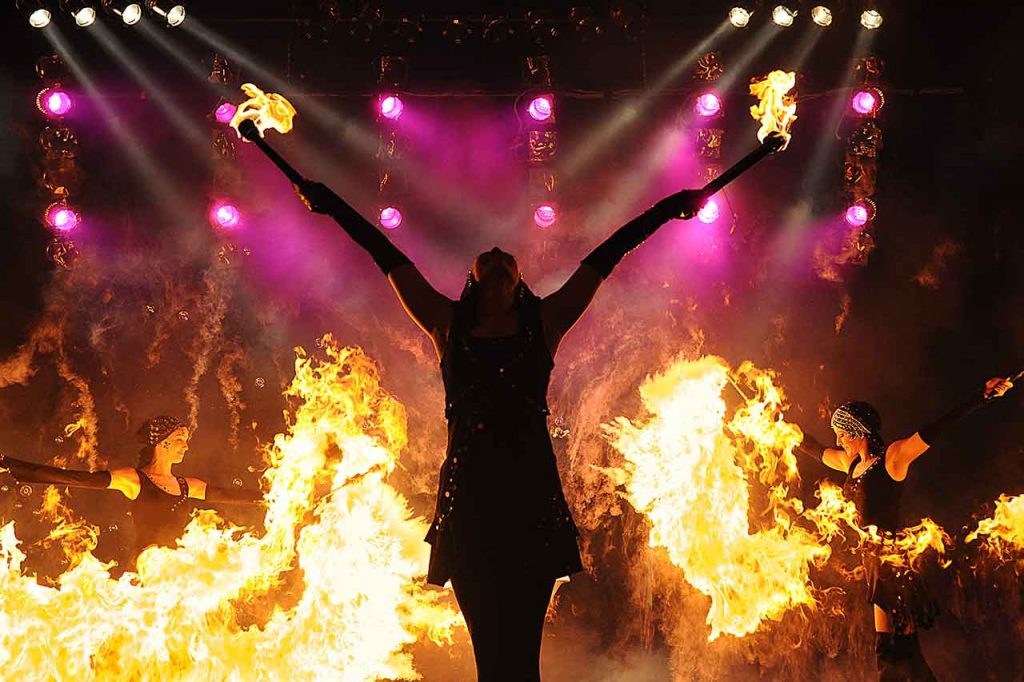 Künstlerin mit 2 Feuerfackeln in den hochgestreckten Armen bei einer Feuershow mit 2 weiteren Backgroundfeuerkünstlerinnen
