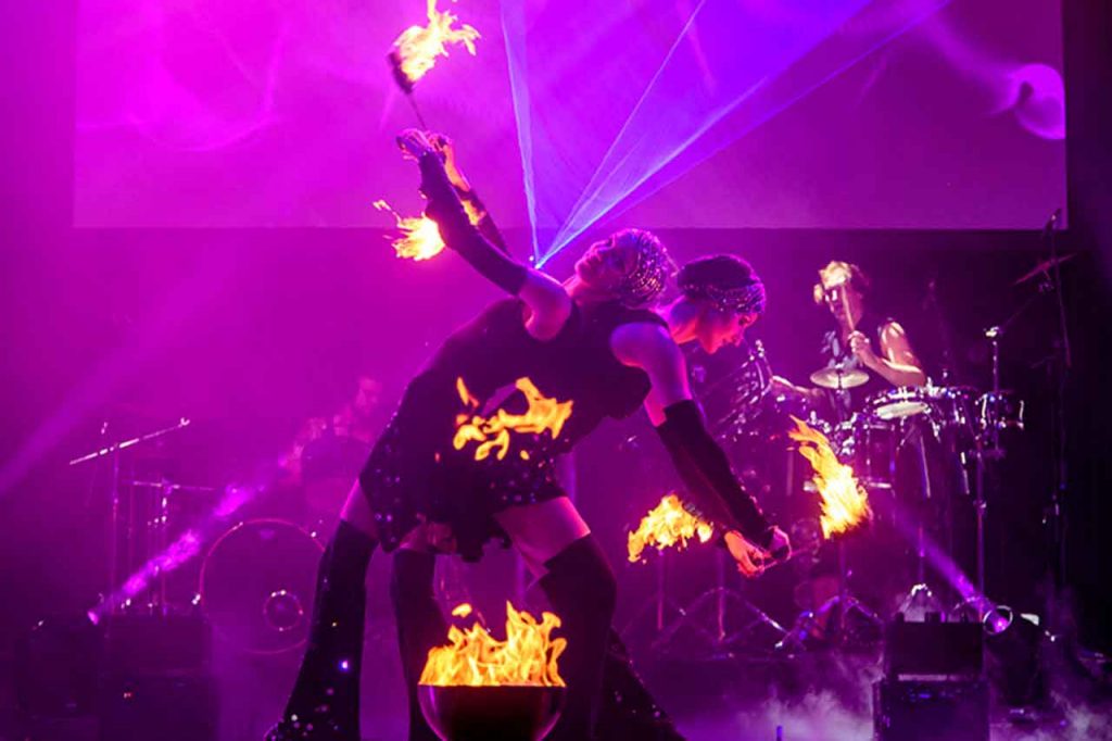 2 Feuerkünstlerinnen mit Feuerfackeln in der Hand und einem Schlagzeuger im Bildhintergrund bei einer FIREDANCER Feuershow